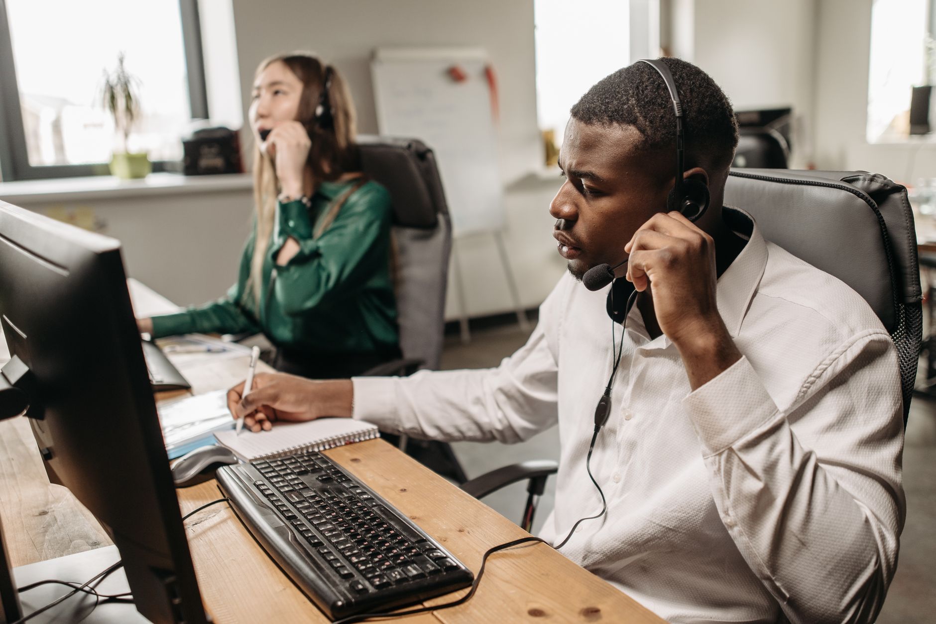 Two office staff wearing headsets at a shared desk, one taking notes while on a call