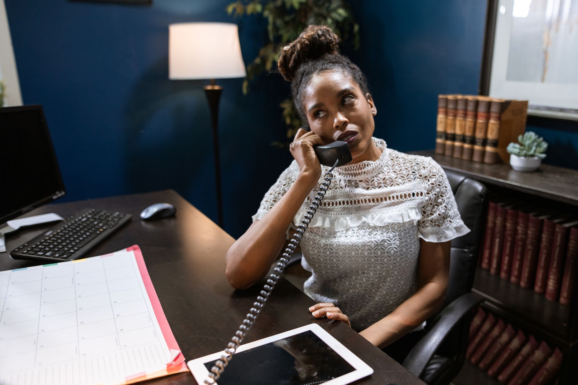 A businesswoman at an office desk takes a call on a landline phone with a desk calendar open in front of her