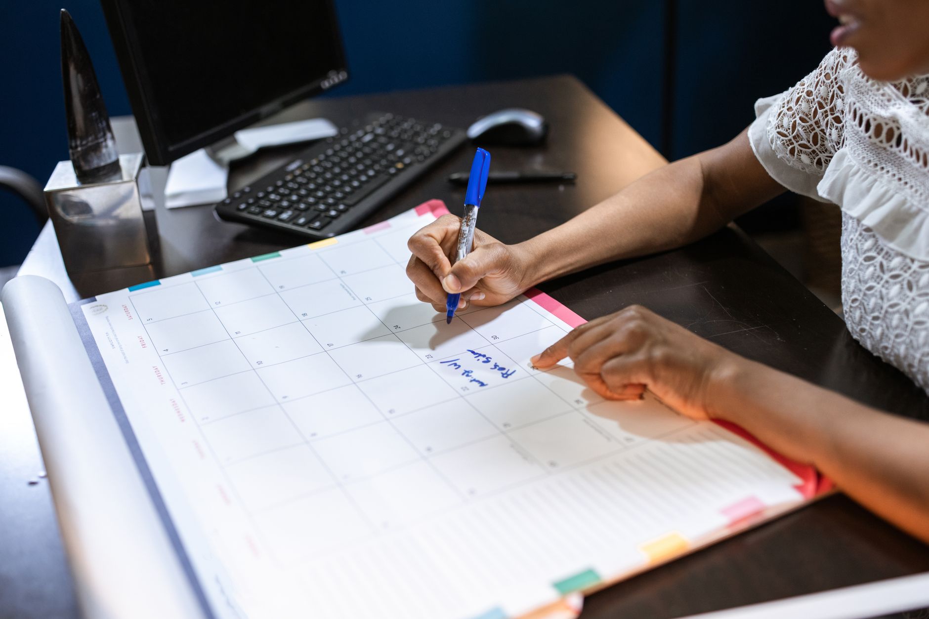 Person writing on a large desk calendar beside a computer keyboard